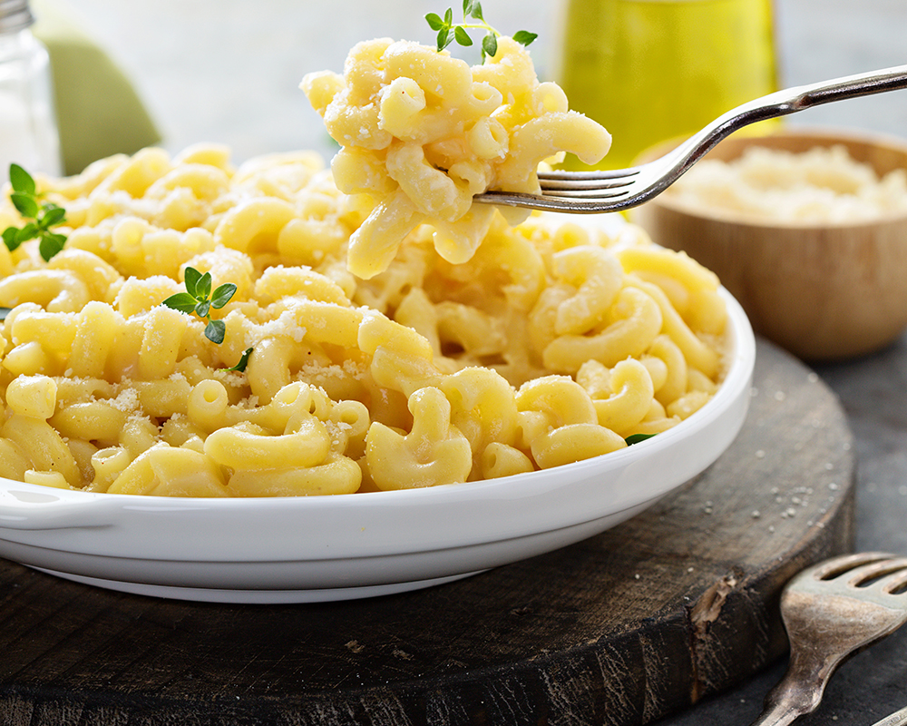 Macarrones con queso en un plato blanco con queso parmesano y especias, sobre una tabla de madera. Alrededor hay cubiertos, un bowl con queso y frasco de aceite