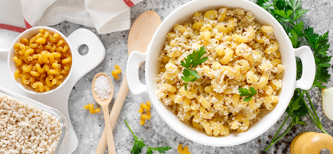 Macarrones con pollo y especias en un plato hondo. Alrededor hay hojas de perejil, utensilios de cocina, un bowl con macarrones crudos y un recipiente con pollo desmenuzado. Todo sobre un fondo gris."