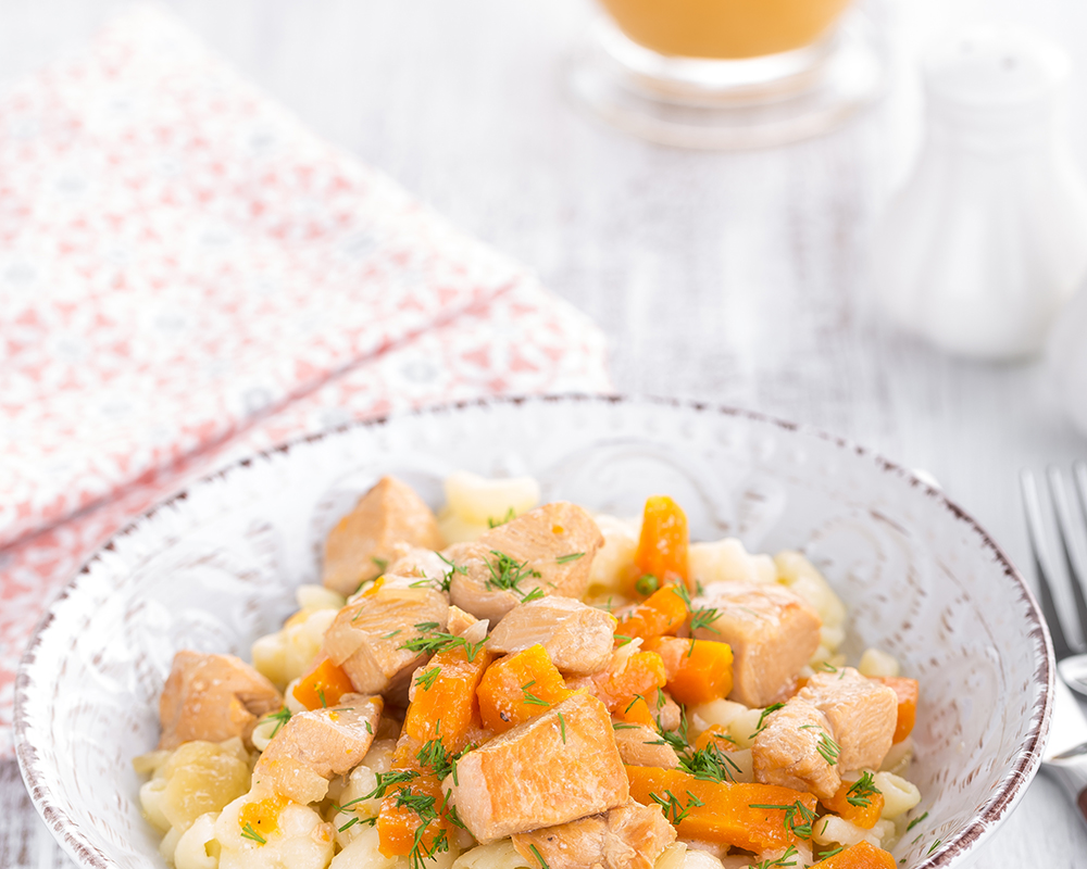 Macarrones con queso y pollo y especias en un plato hondo. Alrededor hay cubiertos, una servilleta de tela y u vaso con jugo. Todo sobre una superficie clara.