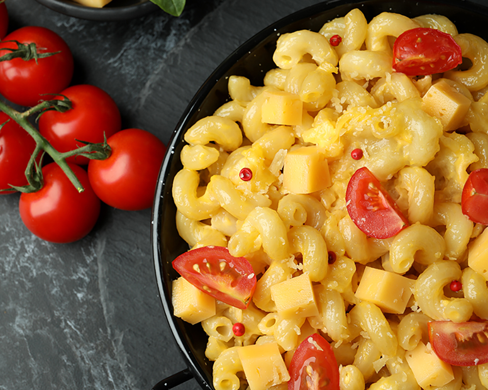 Macarrones con trozos de queso, tomates cherry en un plato negro. Alrededor hay cubiertos, tomates cherry y trozos de queso. Todo sobre un fondo oscuro.