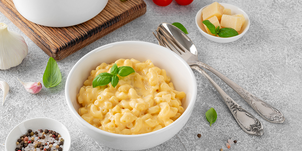 Macarrones con queso en un bowl blanco con albahaca encima sobre un fondo gris. Alrededor hay cubiertos, un bowl de semillas, albahaca y queso en cubos