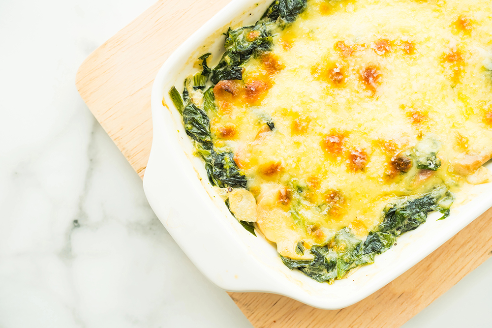 Pasta horneada con espinaca y queso gratinado en una refractaria blanca sobre una tabla de madera. Todo reposa en una superficie de mármol.