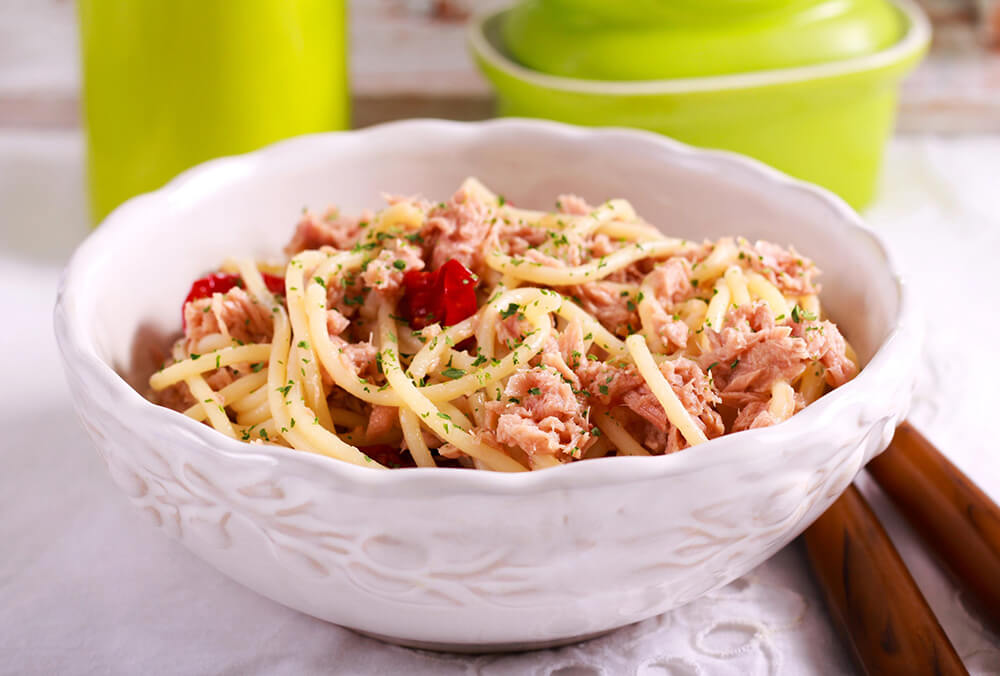 asta Spaghetti con trozos de huevo cocido y tomate, en un bowl blanco. Al fondo se aprecian dos recipientes verdes, todo sobre una superficie blanca.
