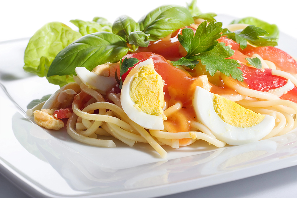 Plato blanco con spaghettis acompañados de huevo cocido en trozos, tomate y albahaca para decorar.