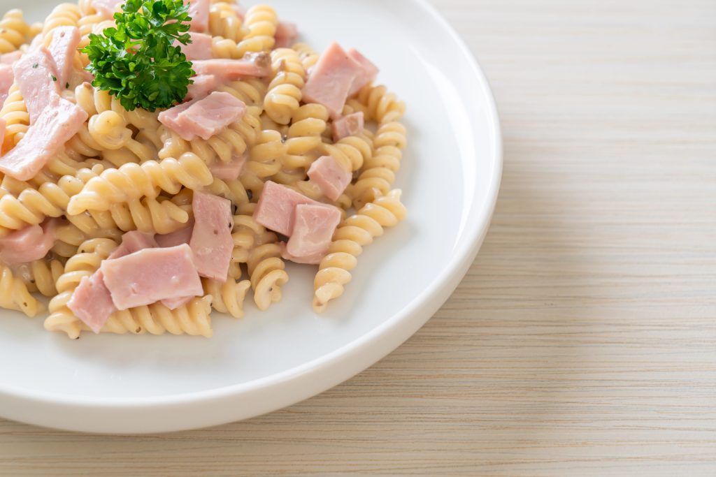 Vista de un plato de pasta tornillo, cremoso con pedazos de jamón y decorado con una rama de cilantro. Todo sobre un plato blanco, que a su vez, está sobre una superficie de madera clara.