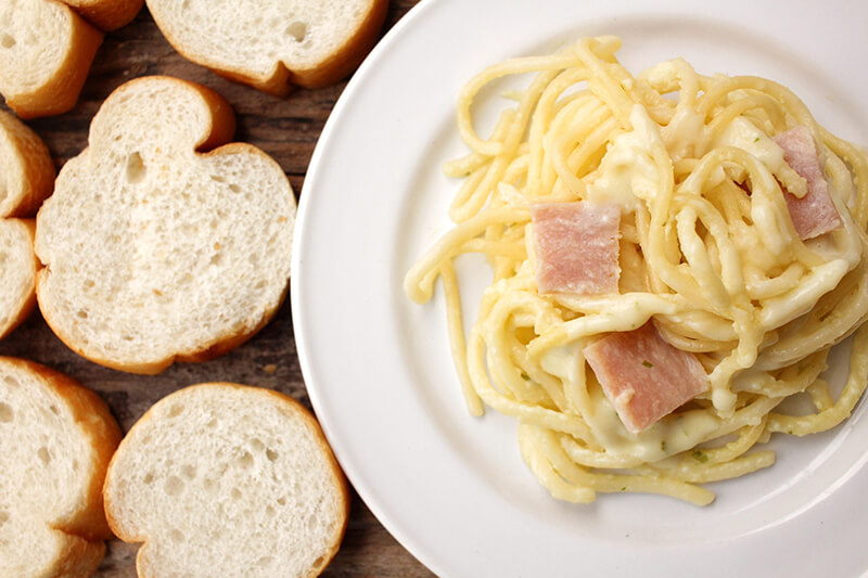 Vista superior de un plato pasta con jamón sobre una superficie de madera oscura. Al lado izquierdo del plato se aprecian trozos de pan francés que lo acompañan.