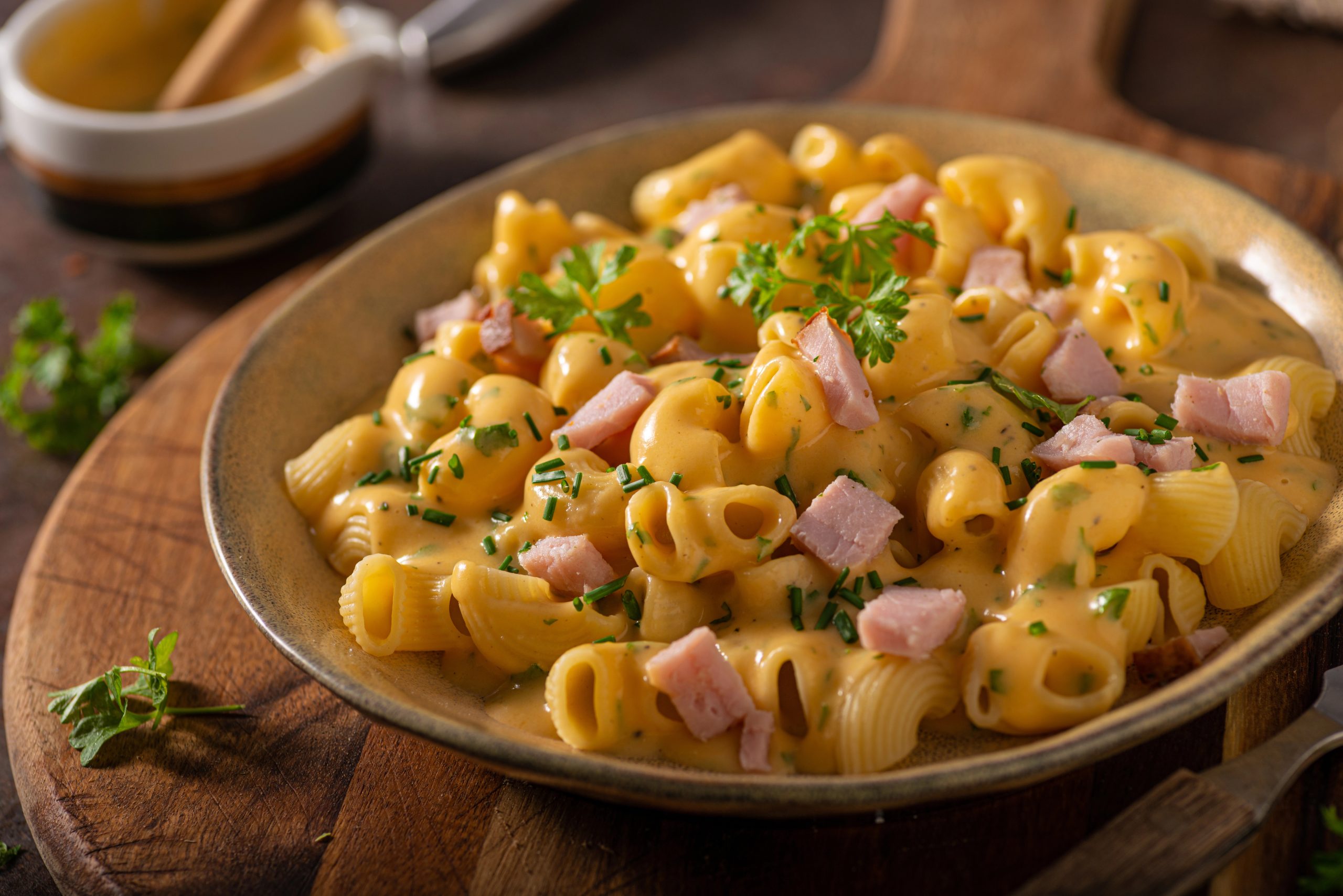 Vista de un plato tipo bowl con macarrones, salsa de queso cheddar y trocitos de jamón, decorado con hojas de cilantro. Todo sobre una bandeja rústica de madera, acompañada por un recipiente con más salsa de queso cheddar