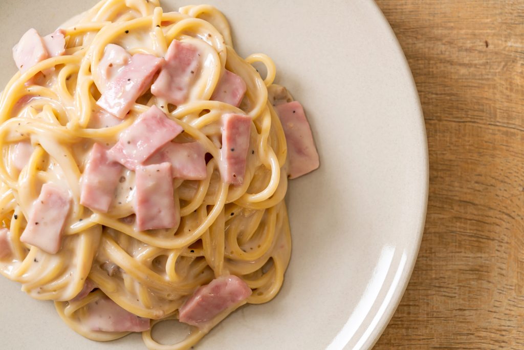Vista superior de un plato de spaghetti cremoso con pedazos de jamón.Todo sobre un plato blanco, que a su vez, está sobre una superficie de madera clara.