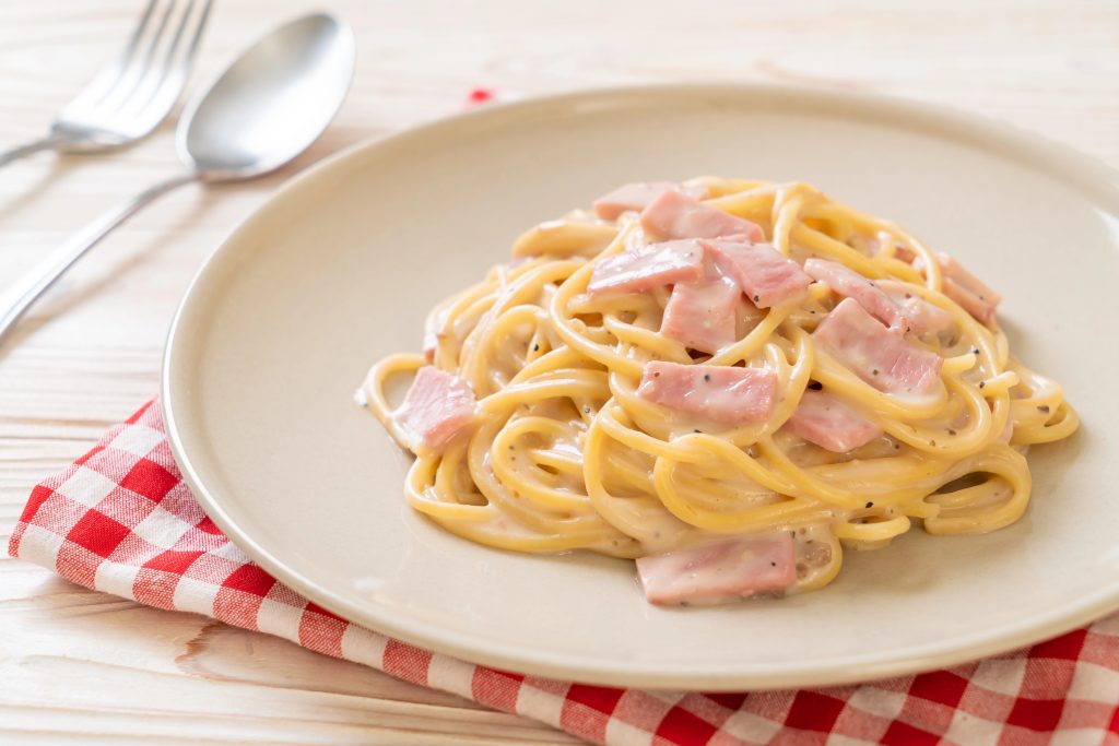 Vista de un plato de spaghetti cremoso con pedazos de jamón.Todo sobre un plato blanco, que a su vez, está sobre una tela a cuadros rojos y blancos y superficie de madera clara. Al lado izquierdo hay un juego de cubiertos.