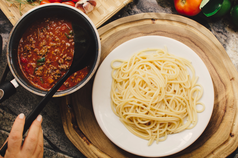 Spaghetti servido en un plato blanco junto a una sartén con salsa boloñesa casera lista para acompañar la pasta