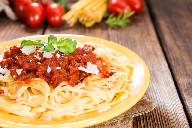 Spaghetti a la boloñesa con salsa de carne y tomate, queso parmesano y hojas de albahaca, servido en un plato amarillo sobre una mesa de madera.