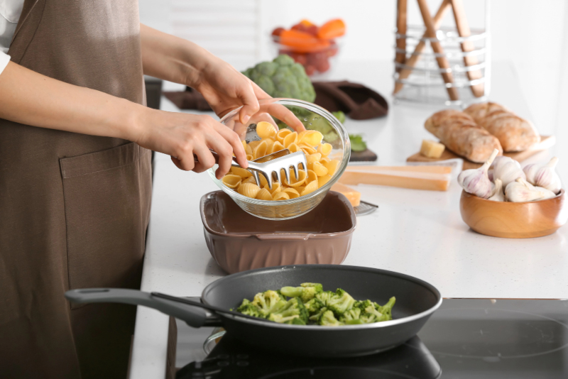 Persona sirviendo pasta corta tipo concha junto a vegetales verdes durante la preparación de una receta en cocina.